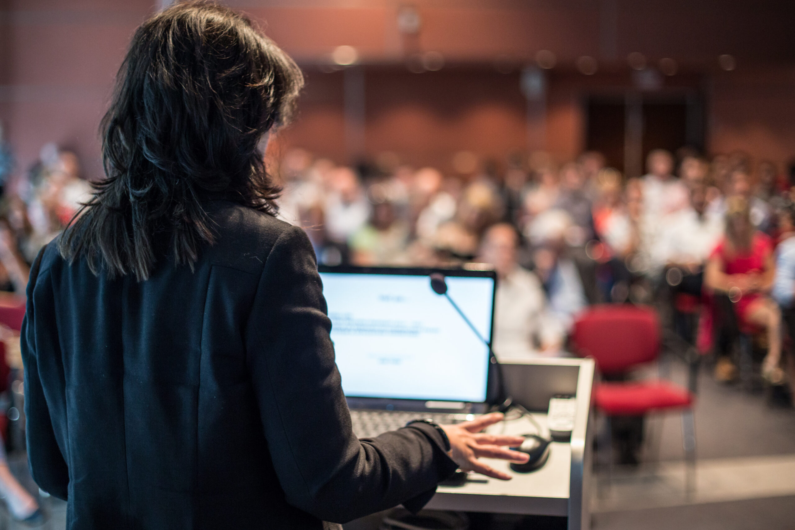 Association professional speaking at a podium to a room full of people at a conference hall.